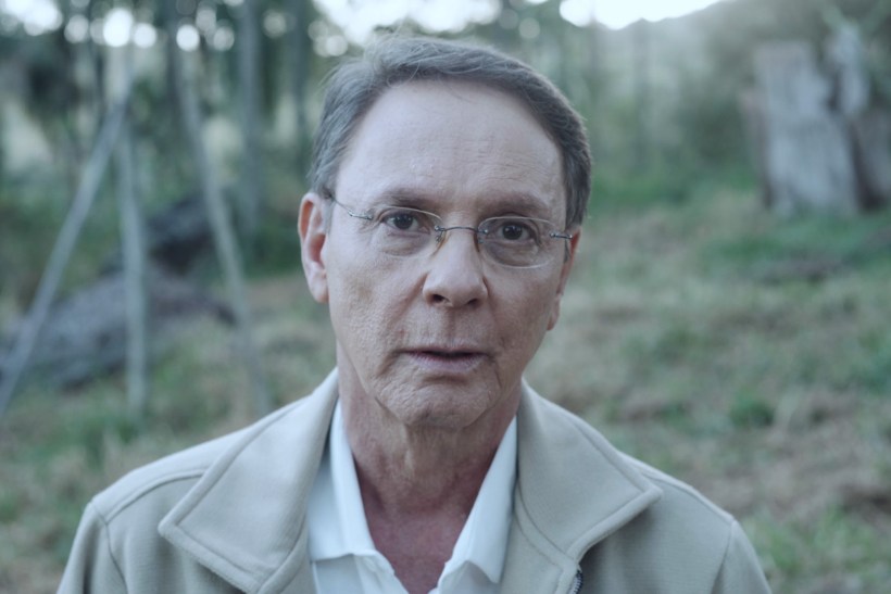 Retrato de um homem de meia-idade, com óculos e uma expressão séria, em um ambiente natural com árvores ao fundo.