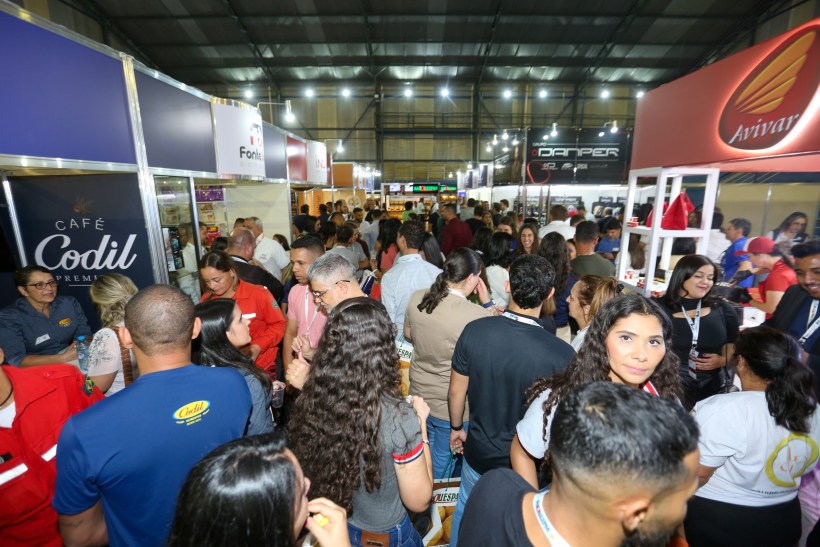 Multidão de pessoas interagindo em um evento de varejo, com estandes de empresas em um ambiente interno destacado por iluminação artificial. Pessoas conversam e trocam informações enquanto exploram produtos e serviços.