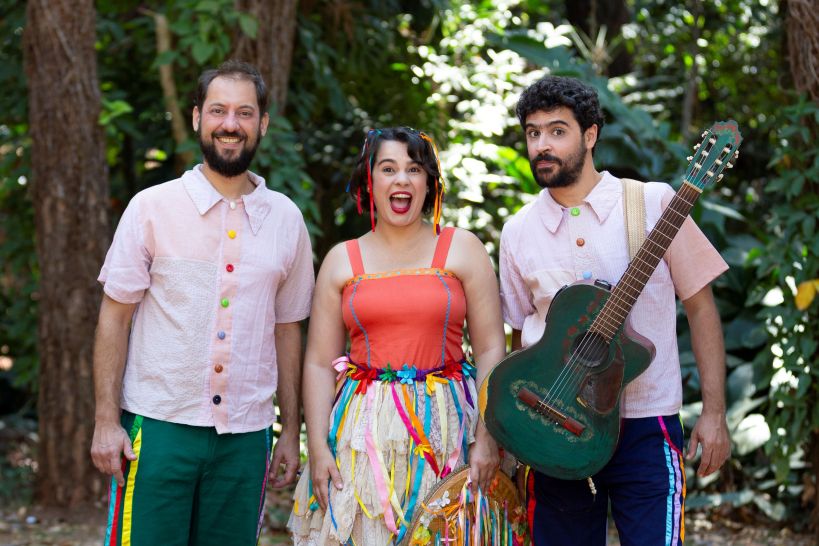 Três integrantes do Grupo Maria Cutia posam juntos em um ambiente natural, usando roupas coloridas e alegres. Um dos membros segura uma guitarra enquanto todos sorriem para a câmera.