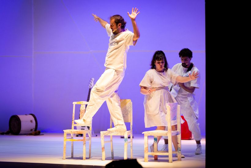 Três atores do Grupo Maria Cutia se apresentam em um palco colorido, com movimento dinâmico e expressões de alegria, enquanto um deles está sobre uma cadeira, em uma encenação teatral. O cenário é iluminado em tons de roxo e branco.