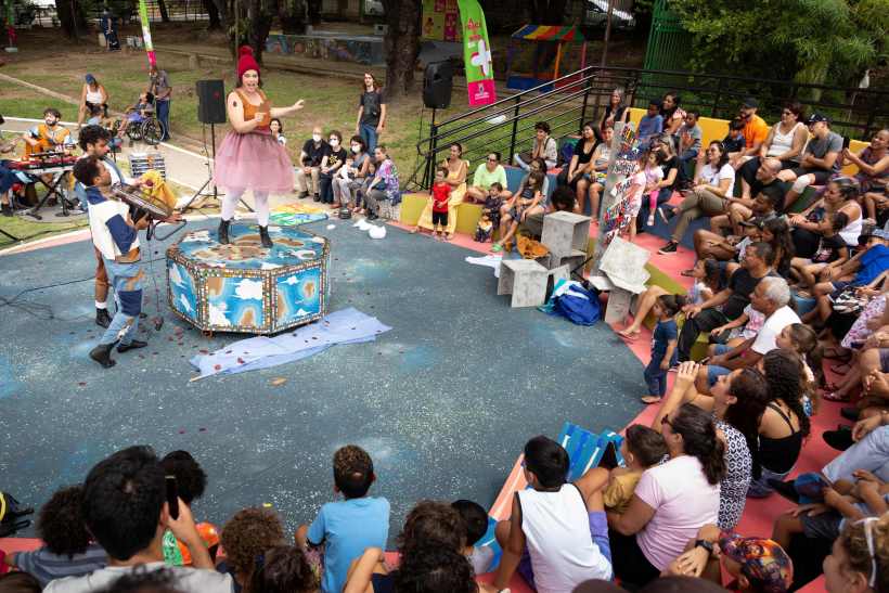 Apresentação do Grupo Maria Cutia em um espaço ao ar livre, com uma performer central vestida de forma lúdica e músicos ao fundo, em frente a um público variado em um ambiente familiar e colorido.
