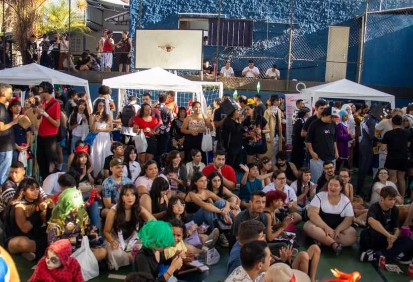 Uma multidão de pessoas vestidas em trajes de cosplay assiste a um evento de cultura pop oriental em um local coberto, com tendas brancas ao fundo.