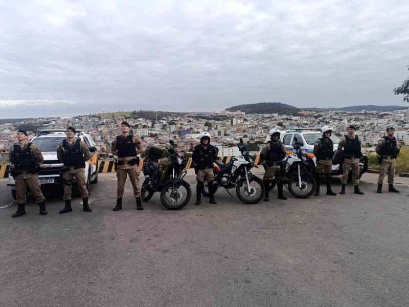Um grupo de policiais militares em motocicletas e viaturas, posando em uma área elevada, com vista da cidade ao fundo.