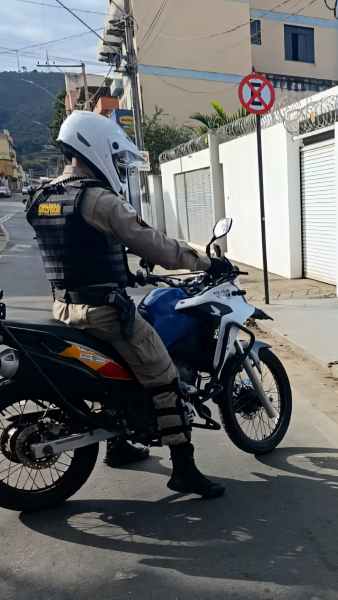 Policial militar pilotando uma motocicleta em uma rua, com uma placa de proibição de estacionamento ao fundo.