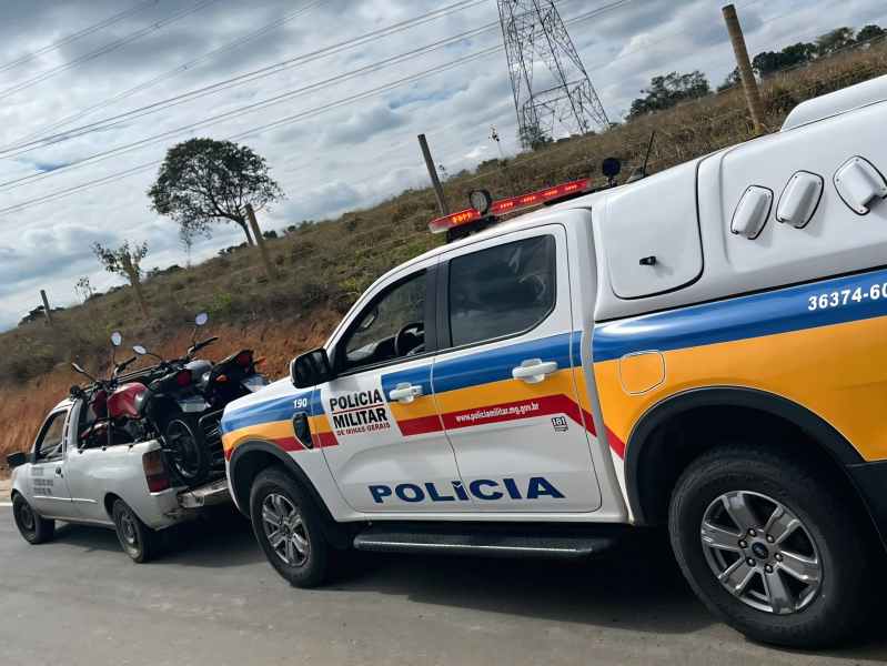 Uma viatura da Polícia Militar de Minas Gerais estacionada ao lado de um carro que transporta motocicletas apreendidas, em uma estrada com vegetação ao fundo.
