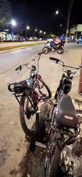 Duas bicicletas estacionadas na calçada à noite, com uma motocicleta ao fundo e iluminação pública visível.