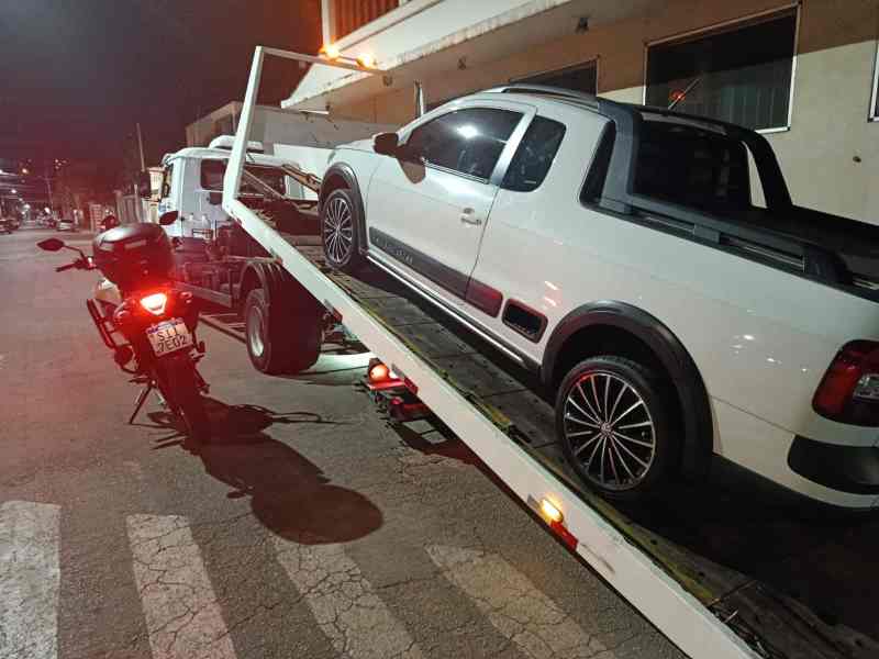 Caminhão guincho removendo um veículo em uma rua à noite, com uma moto estacionada ao lado.