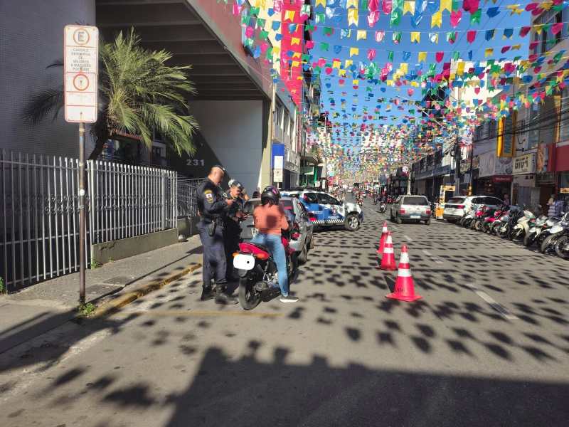 Guarda Civil Municipal realizando uma abordagem em motociclista durante a Operação Impacto em área urbana decorada com bandeirinhas coloridas.