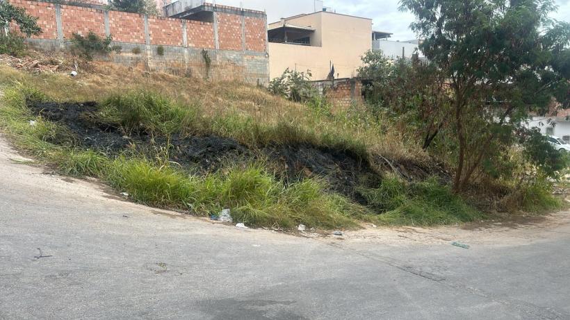Vista de um terreno com vegetação queimada e grama alta em um bairro urbano, com resíduos visíveis no chão.