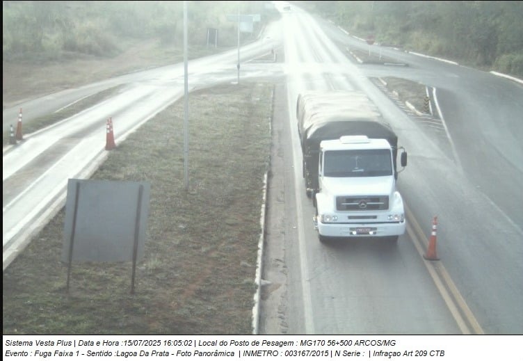 Imagem de uma rodovia com um caminhão branco transportando carga, registrado em um posto de pesagem na MG170, em Arcos/MG, com cones de sinalização visíveis e um painel de trânsito ao fundo.