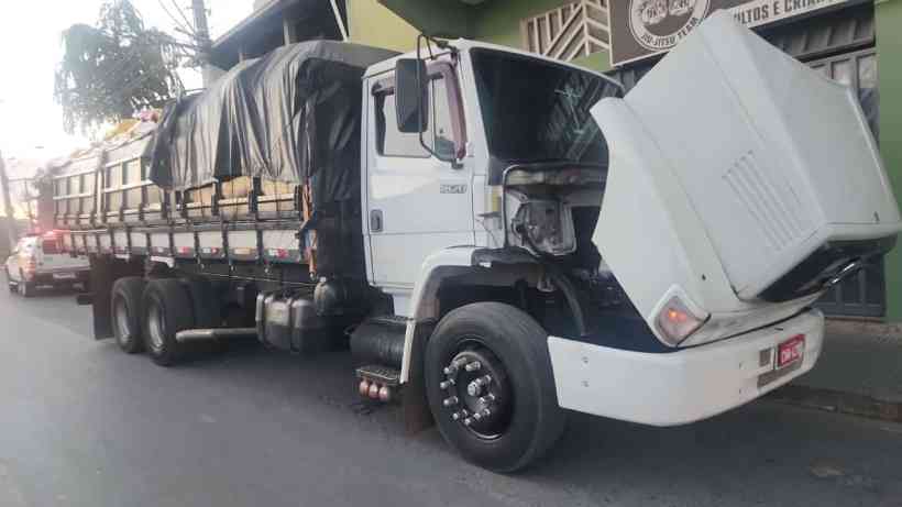 Caminhão M.Benz branco com a cabine levantada, estacionado em uma rua. O veículo está carregado e coberto com uma lona, apresentando placas de identificação parcialmente ocultas.