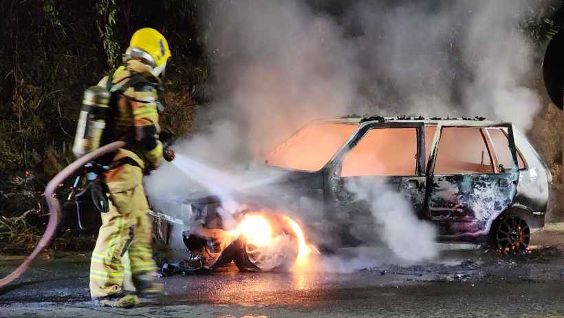 Um bombeiro combatendo um incêndio em um veículo Fiat Uno, ano 1995, que está em chamas na rodovia Carmem Duarte, com fumaça e fogo visíveis.