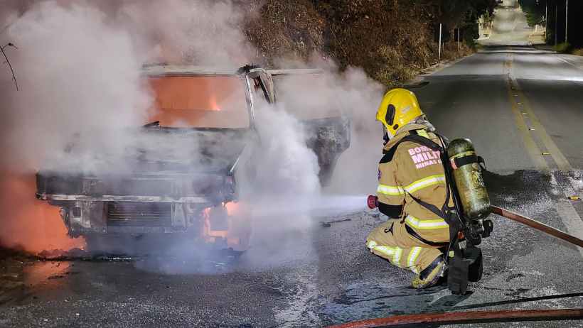 Um bombeiro combatendo as chamas de um veículo Fiat Uno em chamas na rodovia Carmem Duarte, acompanhado de fumaça e material de combate a incêndios.