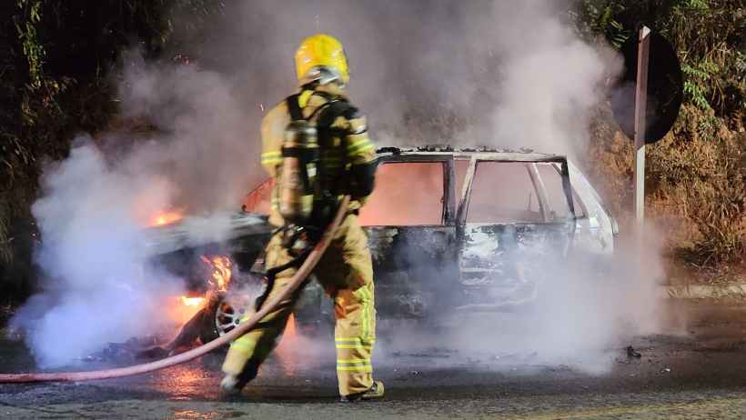 Bombeiros apagando incêndio em veículo Fiat Uno em Nova Serrana, com fumaça e chamas visíveis.