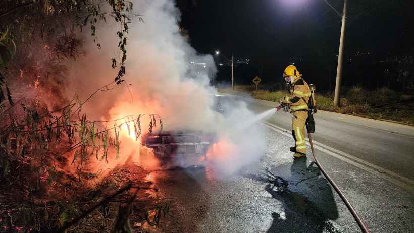 Bombeiro combatendo incêndio em um veículo em uma rodovia à noite, com fumaça e chamas visíveis.