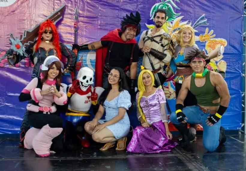 Grupo de pessoas vestidas em trajes de cosplay, posando para a foto em um evento de cultura pop, com um fundo colorido que apresenta personagens icônicos.