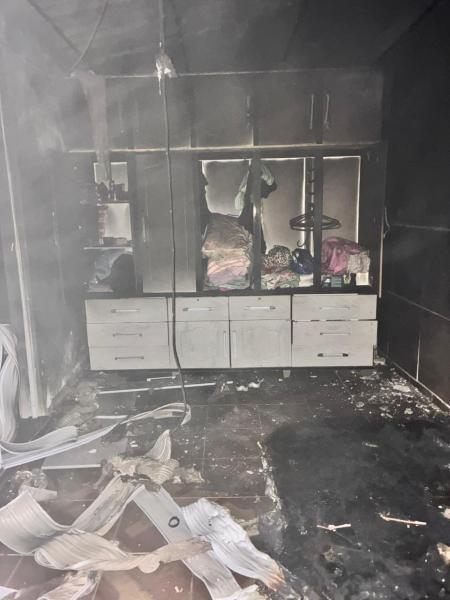 Interior de uma casa danificada por incêndio, com mobília e roupas queimadas, e sinais de fumaça visíveis nas paredes.