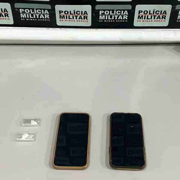 Dois celulares e dois papelotes de substância semelhante à cocaína em uma mesa, com fundo de cartazes da Polícia Militar de Minas Gerais.
