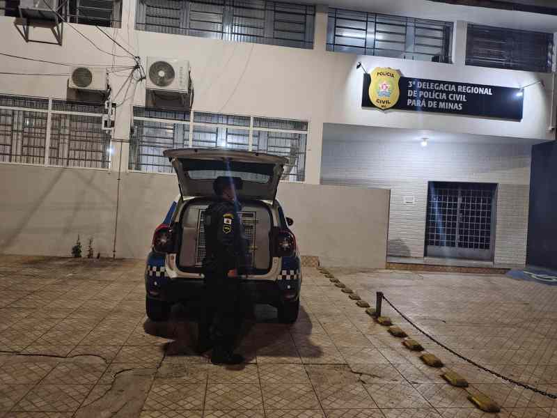 Um agente da Guarda Civil Municipal ao lado de um veículo estacionado em frente à delegacia regional de Polícia Civil em Pará de Minas à noite.