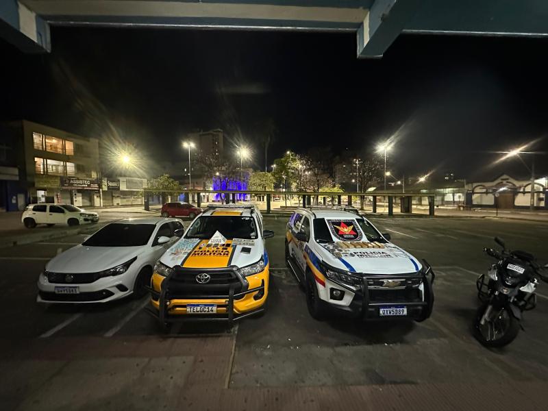 Veículos da polícia militar estacionados em um local iluminado durante a noite, incluindo uma picape e um carro branco à esquerda.