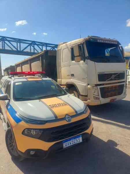 Um veículo da polícia militar estacionado ao lado de uma carreta na rodovia MG 170, com um viaduto visível ao fundo.