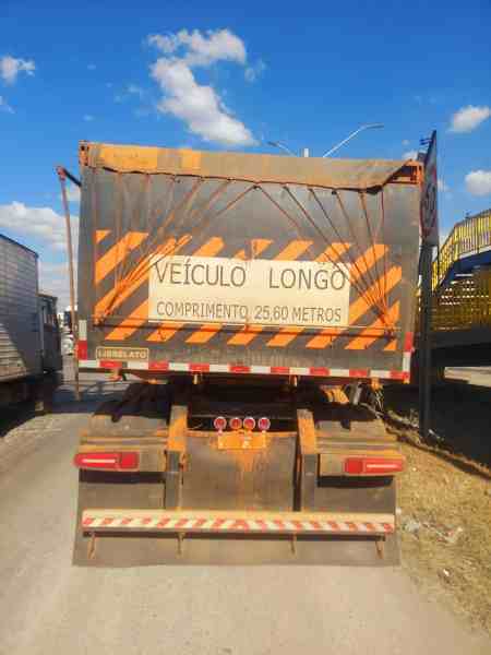 Imagem de um caminhão com sinalização de 'VEÍCULO LONGO' e medidas de comprimento de 25,60 metros, estacionado em uma rodovia sob céu azul com algumas nuvens.