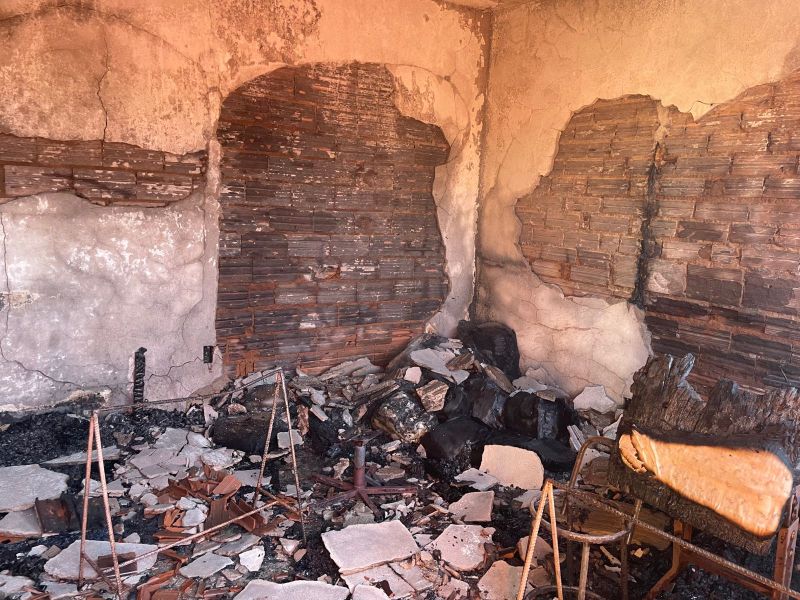 Imagem de uma sala com paredes danificadas por incêndio, mostrando escombros no chão e um sofá parcialmente queimado.
