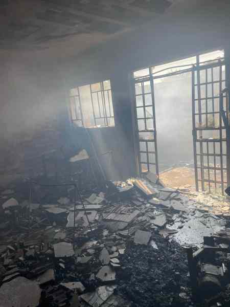 Interior de uma residência devastada por incêndio, com fumaça visível e destroços espalhados pelo chão.