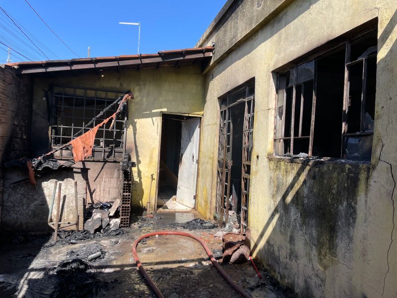 Exterior de uma residência afetada por incêndio, com paredes danificadas, janelas quebradas e restos de materiais queimados. Um mangueira de combate ao incêndio está presente no chão.