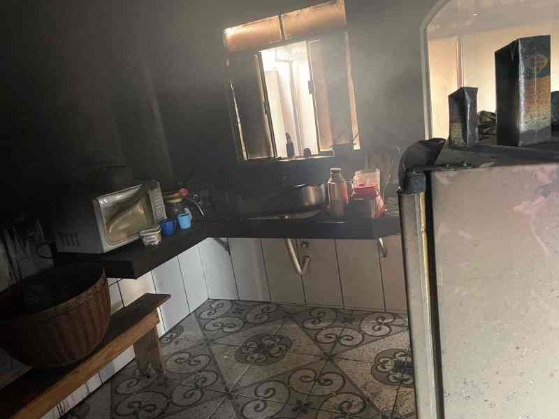 Cozinha de uma residência com fumaça densa, móveis e utensílios visíveis, mostrando os efeitos de um incêndio.