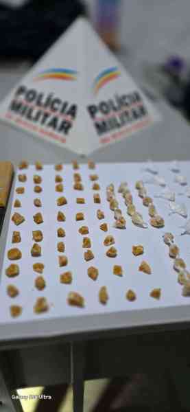Apreensão de drogas, incluindo pedras de crack e porções de cocaína, com placas da Polícia Militar ao fundo.