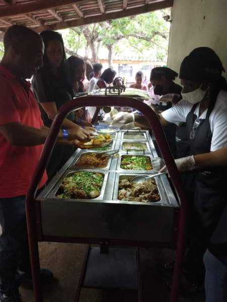 Pessoas servindo e recebendo comida em um evento comunitário, com pratos variados expostos em um carrinho de buffet.
