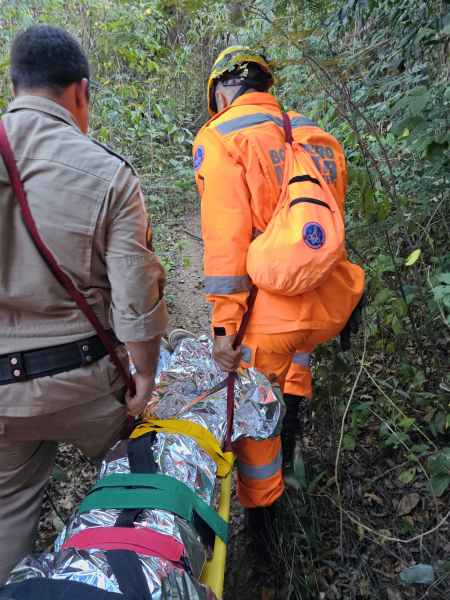 Equipe de resgate levando um idoso imobilizado em uma área de mata fechada, com um bombeiro vestido de laranja e um policial atrás.