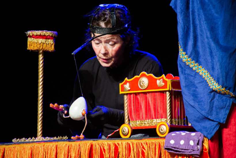 Uma artista de teatro de sombras manipula um ovo em uma mesa colorida, enquanto um pequeno cenário teatral está montado ao fundo.