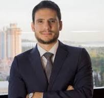 Retrato de um homem em traje formal, com camisa branca e gravata, sorrindo para a câmera em um ambiente de escritório moderno.