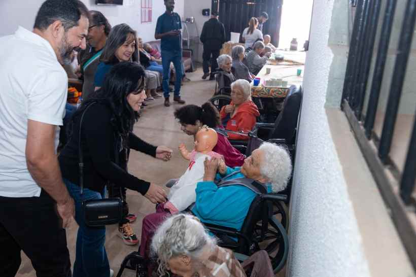 Visita a um lar de idosos com interação afetiva, onde adultos conversam e brincam com os idosos assistidos, incluindo uma mulher segurando uma boneca e vários idosos em cadeiras de rodas.