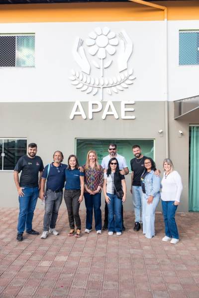 Grupo de pessoas posando em frente à sede da APAAE, com o logotipo da instituição ao fundo.