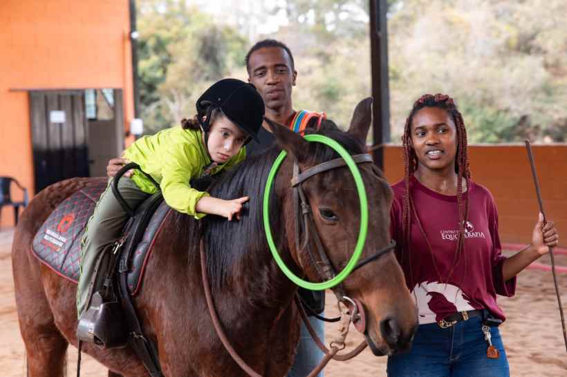 Criança montada em um cavalo, usando um capacete, enquanto é assistida por dois adultos em um ambiente de equoterapia.