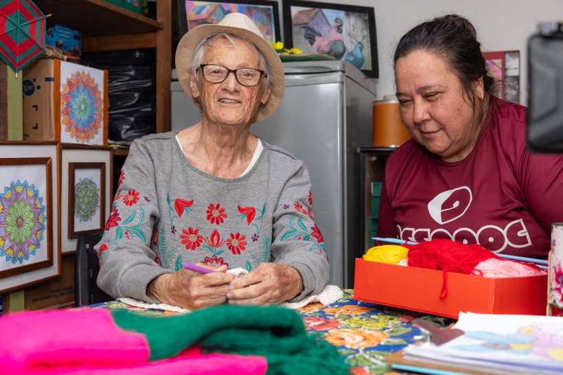 Uma idosa sorridente, usando óculos e um chapéu, está sentada à mesa com uma mulher ao lado, ambas envolvidas em atividades de artesanato. O ambiente é decorado com quadros coloridos e materiais de costura.