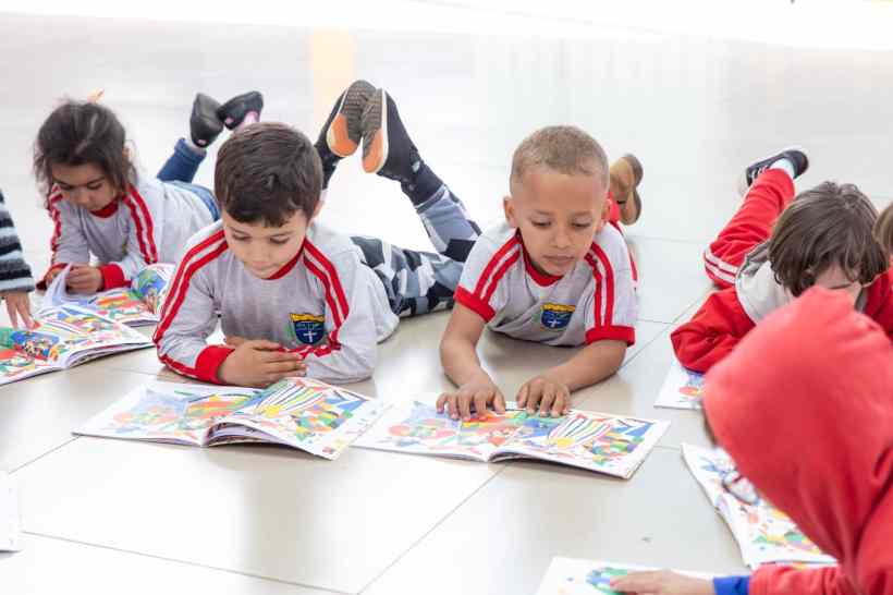 Crianças deitadas no chão lendo livros ilustrados, concentradas em suas atividades, várias vestindo camisetas brancas com detalhes em vermelho.