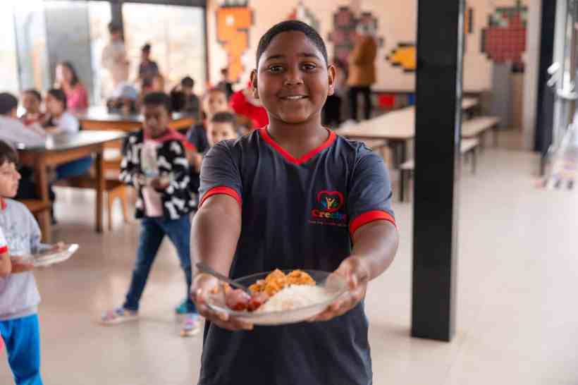Criança sorridente segurando um prato de comida em uma creche, com outras crianças ao fundo.
