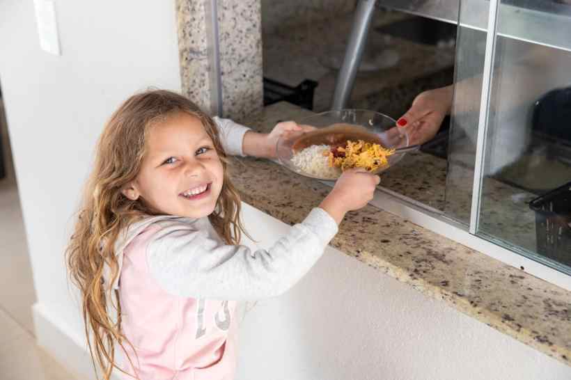 Criança sorridente recebendo comida em um balcão, com uma mão adulta passando um prato com arroz e guarnição.