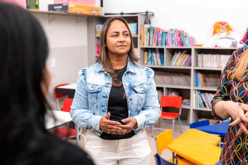 Mulher conversando em uma sala de aula com prateleiras de livros ao fundo.