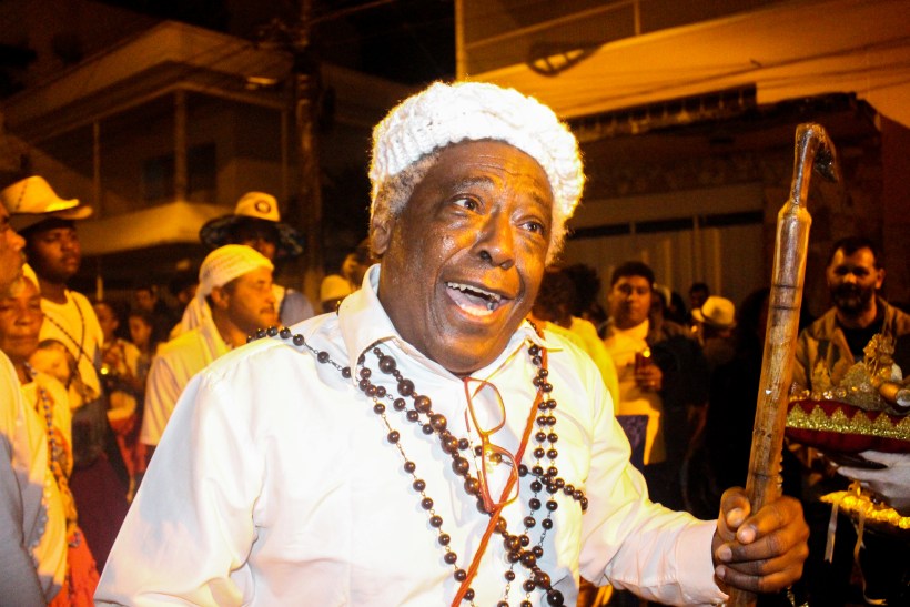 Homem sorridente com vestimenta tradicional, adornado com colares e chapéu, participando de um festival cultural ao ar livre, cercado por outras pessoas.