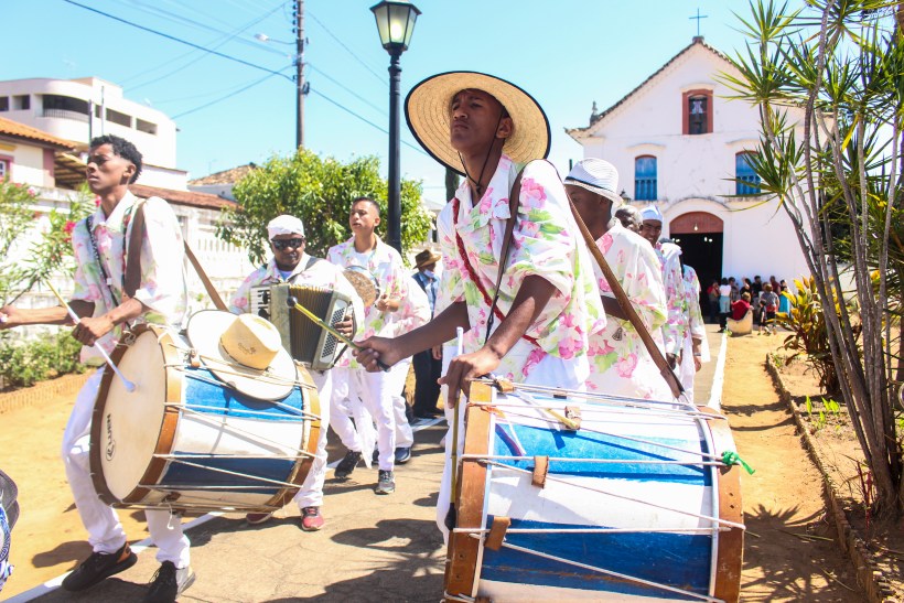 Grupo de músicos e dançarinos com roupas floridas e chapéus, tocando instrumentos de percussão e acordeão durante o Grande Reinado do Rosário em Itapecerica.