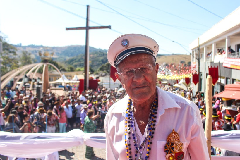 Um homem idoso, usando um chapéu branco e óculos, está em primeiro plano com uma multidão festiva ao fundo durante o Grande Reinado do Rosário em Itapecerica.