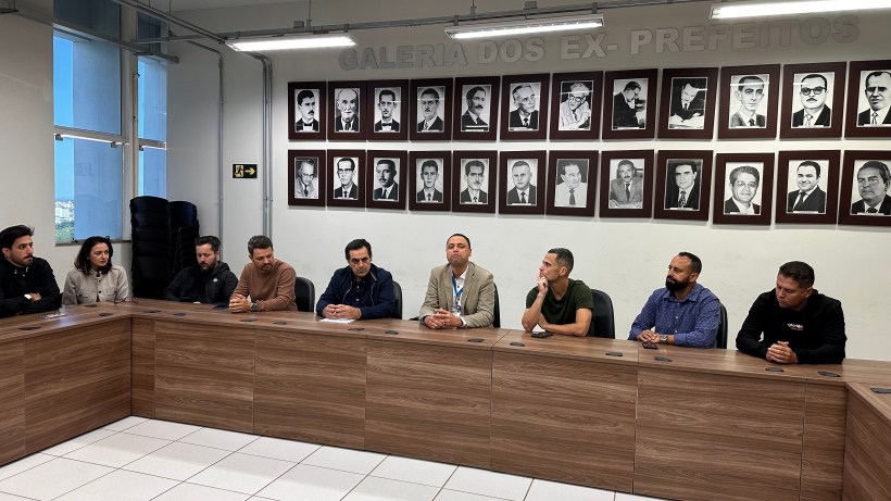 Reunião de representantes em uma mesa, com retratos de ex-prefeitos ao fundo.