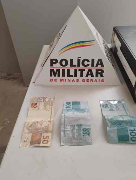 Notas de dinheiro, incluindo cédulas de 50 e 100 reais, dispostas ao lado de um objeto em forma de pirâmide com a inscrição 'POLÍCIA MILITAR DE MINAS GERAIS'.