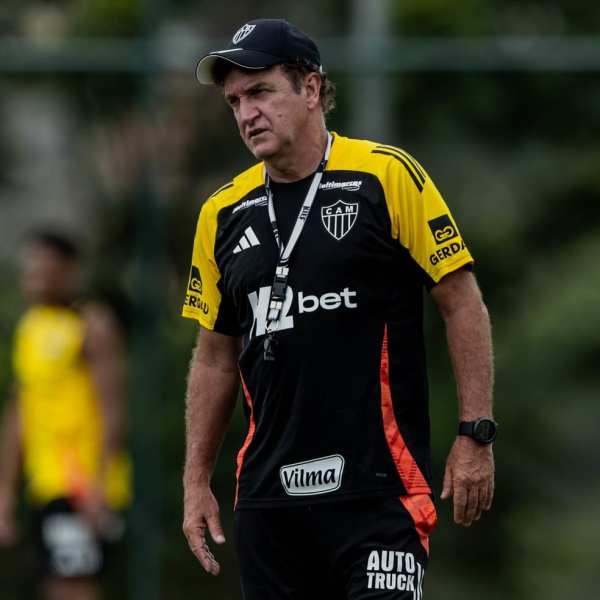 Treinador do Atlético-MG em campo, com uniforme preto e amarelo, observando os jogadores durante o treinamento.