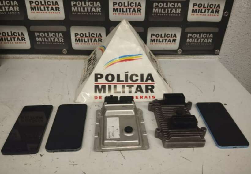 Objetos apreendidos pela Polícia Militar, incluindo quatro aparelhos celulares e duas centralinas veiculares, dispostos sobre uma mesa com fundo de parede da polícia.